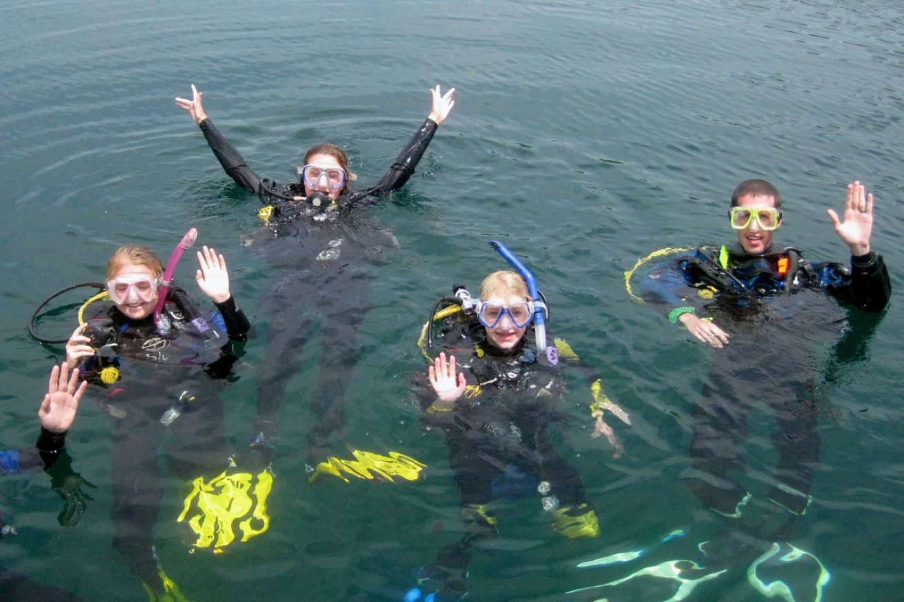 Teens smiling and waving in scuba gear on the surface of the water