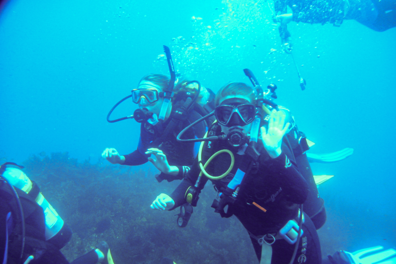 Two female teen scuba divers in the water waving at the camera
