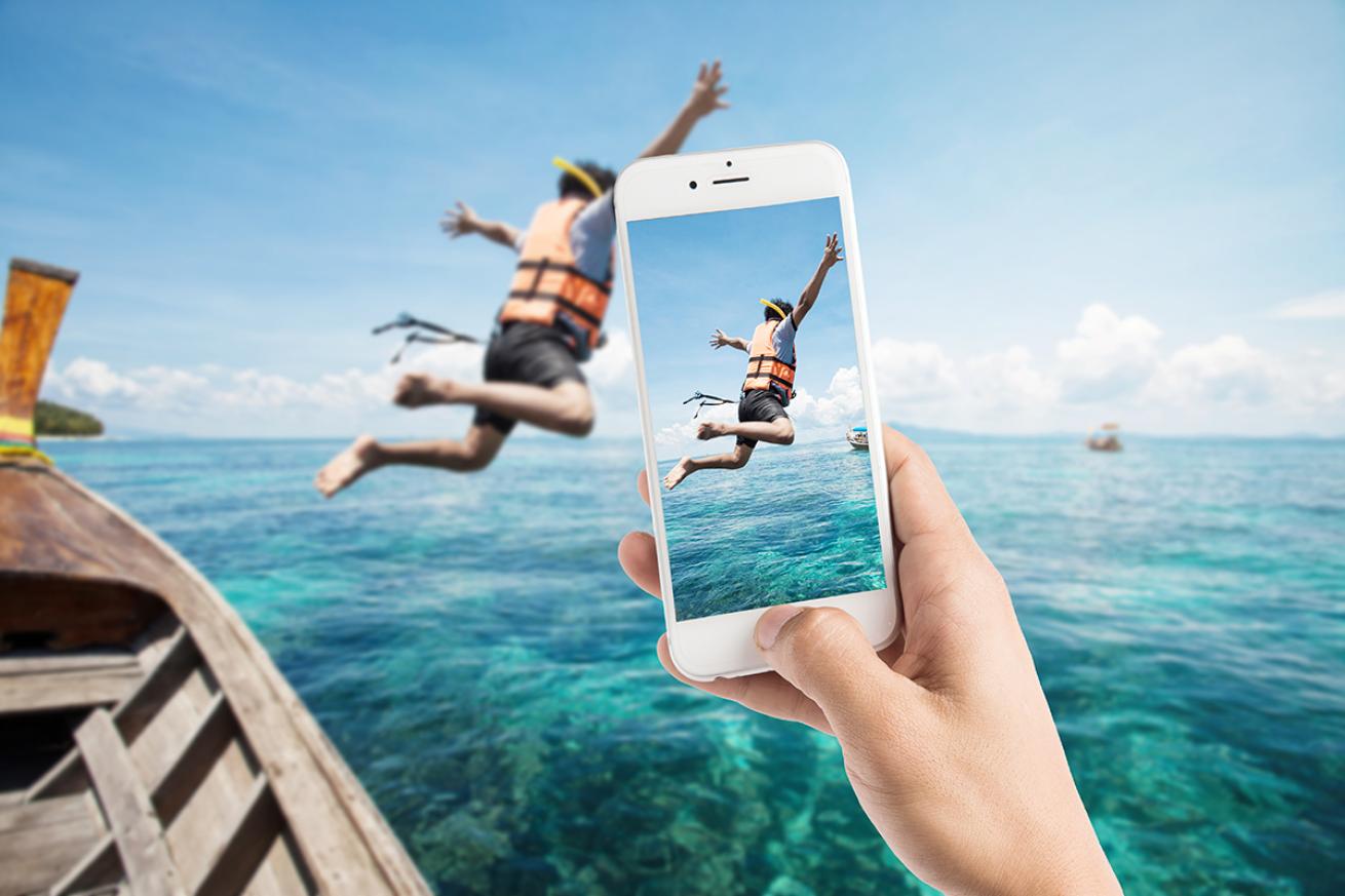 Person taking photo on phone of kid wearing a lifejacket while jumping off boat.