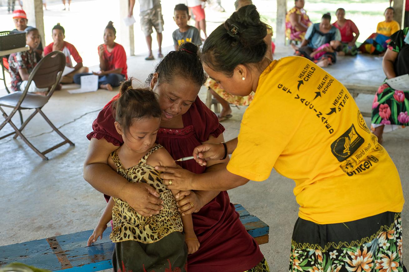 Young girl gets a shot from a female health worker while sitting on her mom's lap/